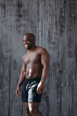 Çıplak gövdeli Afro-Amerikalı genç kaslı adam pazılarını gösteriyor. Gri bir arka planda güneşte duruyor