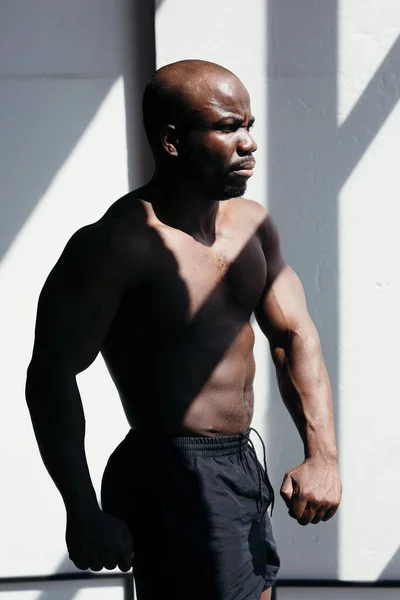 Hombre afroamericano serio entrenador de fitness demuestra su figura a la luz del sol sobre un fondo blanco — Foto de Stock