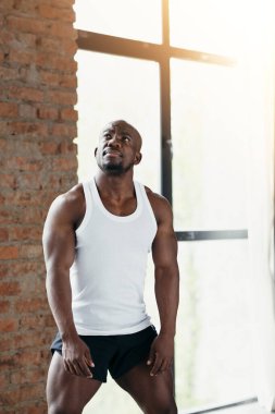Beyaz tişörtlü ve şortlu kaslı erkek vücut geliştirici panoramik pencerenin önünde duruyor ve kaslarını gösteriyor..