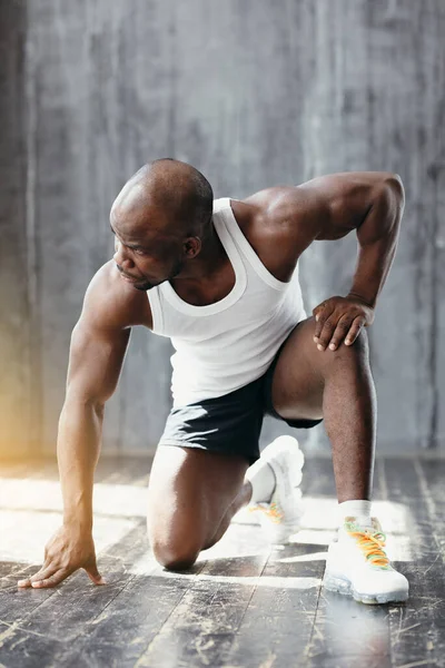 Sports shooting of a muscular dark-skinned trainer in a white T-shirt crouched in a pose to start running in the gym.