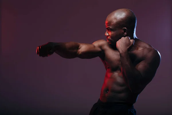 Tmavý boxer trénuje s nahým trupem. Sportovec s reliéfními svaly je podsvícen červeným světlem. — Stock fotografie