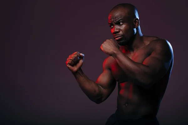 Un afroamericano atlético de pecho desnudo se para en una pose de boxeador para comenzar los ejercicios con un saco de boxeo en una luz de neón . — Foto de Stock