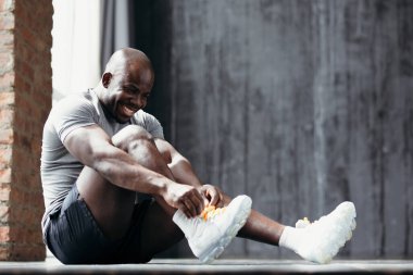 Gri tişörtlü kaslı erkek vücut geliştirici spor salonunda yerde otururken spor ayakkabısının bağcıklarını bağlıyor.