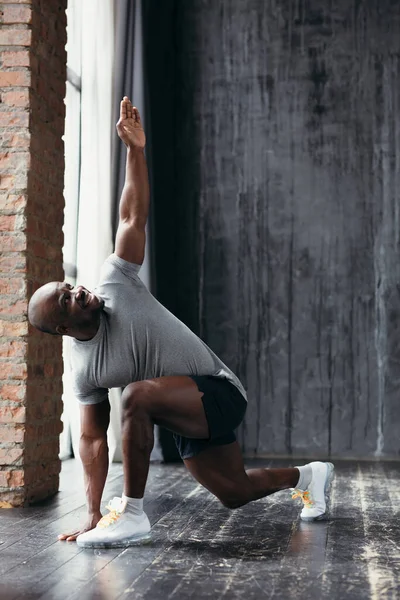 Dark-skinned Pilates trainer in a gray T-shirt doing lunges forward exercise with raising his left hand up