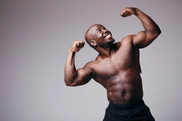 Boxer masculino africano demonstra bíceps e solavancos. Atleta sorri em um fundo cinza — Fotografia de Stock