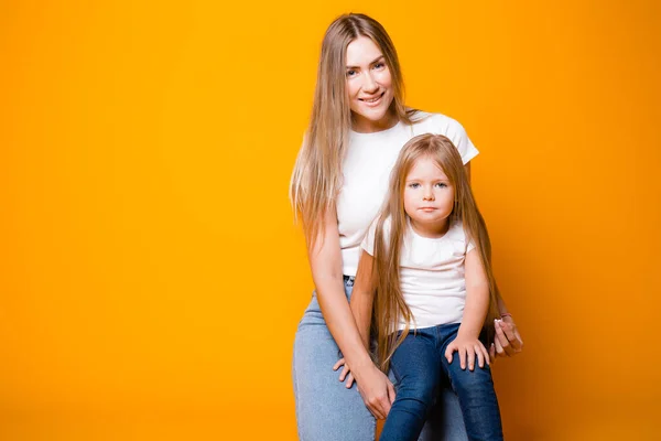 Vacker mor och dotter i vita t-shirts och blå jeans sitta på en orange bakgrund — Stockfoto