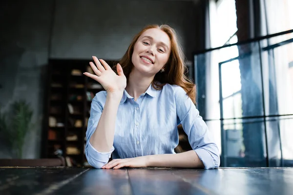 Une Américaine en chemise bleue s'assoit au bureau, agite la main en saluant et sourit à la caméra — Photo