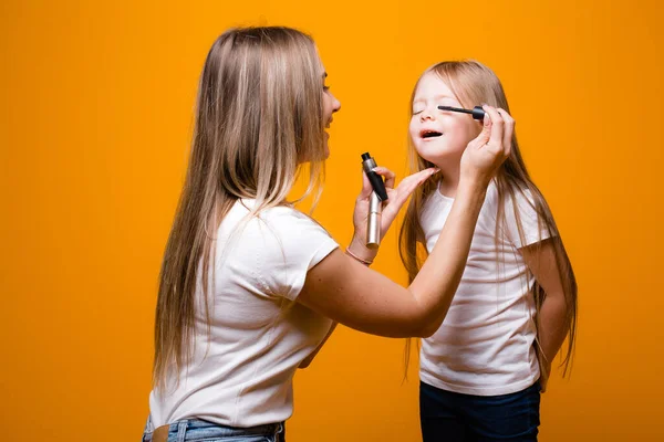 Мама делает макияж для глаз своей дочери. Краски туши на оранжевом фоне — стоковое фото