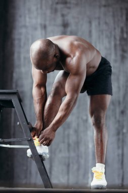 Çıplak gövdesi ve beyaz spor ayakkabıları olan esmer tenli bir adam ayağını sandalyeye dayayıp ayakkabı bağcıklarını bağlıyor.