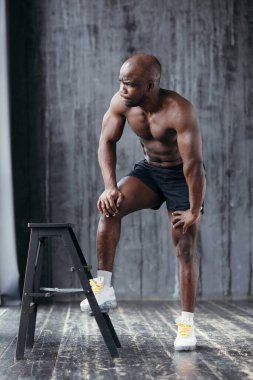 Koyu tenli, çıplak gövdeli, beyaz ayakkabılı bir adam ayağını sandalyeye koyup stüdyonun penceresinden dışarı bakıyor.