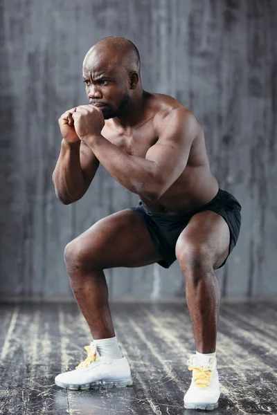 Athletic goły klatka piersiowa Afroamerykanin robi przysiady trzymając się za ręce w pobliżu klatki piersiowej w siłowni — Zdjęcie stockowe