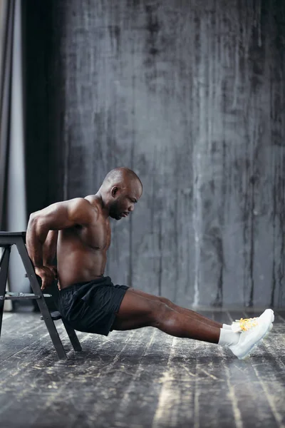 Visualização de perfil de afro-americano com tronco nu e tênis brancos apoiados em uma cadeira e empurrando para cima balança os músculos do braço — Fotografia de Stock
