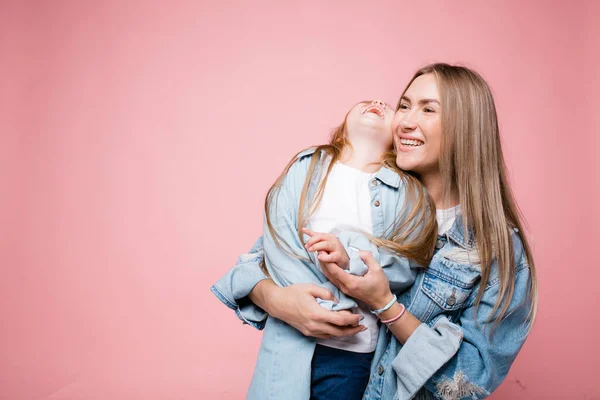 Långhåriga mor kramar sin dotter hårt, medan hon kastade huvudet tillbaka i skratt. Lycklig familj på en rosa bakgrund — Stockfoto