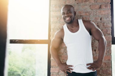 Beyaz tişörtlü ve siyah şortlu Afro-Amerikan portresi kameranın önünde spor salonunun arka planında poz veriyor..