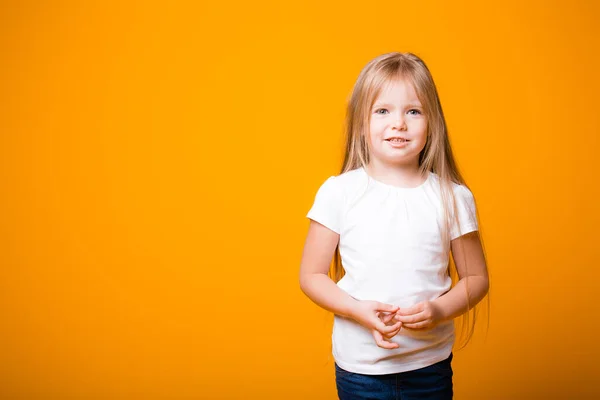 Blond haar meisje kind staat in een wit T-shirt en jeans op een oranje achtergrond en glimlacht — Stockfoto