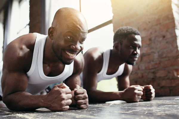 Dwóch Afroamerykanów w białych koszulkach trenuje mięśnie brzucha i pleców stojąc z przedramionami i nogami na podłodze.. — Zdjęcie stockowe