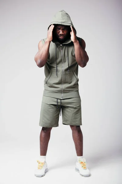 A strong and muscular African American in white crosses pulls the hood of a sweatshirt over his face, demonstrating a tracksuit.