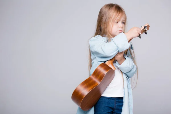 The child tunes the guitar. Girl learning to play ukulele music. Posing on a gray background with ukulele