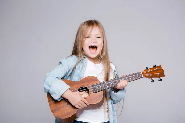 Das Mädchen singt und spielt Ukulele. Das Kind lacht, posiert für die Kamera und genießt die Musik — Stockfoto