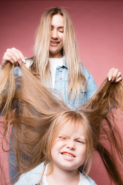 Mama próbuje rozplątać długie blond włosy córki. Dziecko nie chce iść do fryzjera. Izolacja na różowym tle — Zdjęcie stockowe