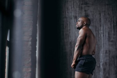 Güçlü ve kaslı Afro-Amerikalı çıplak bluzu ve siyah şortu ile pencereden dışarı düşünceli bir şekilde bakıyor.
