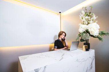 Bir kliniğin modern resepsiyonunda çalışan genç ve güzel bir kadın bilgisayardan randevu alırken telefona cevap veriyor.