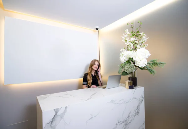 Modern klinikte genç resepsiyonist telefonda konuşuyor. Arkaplanda kopyalama alanı olan boş beyaz poster.