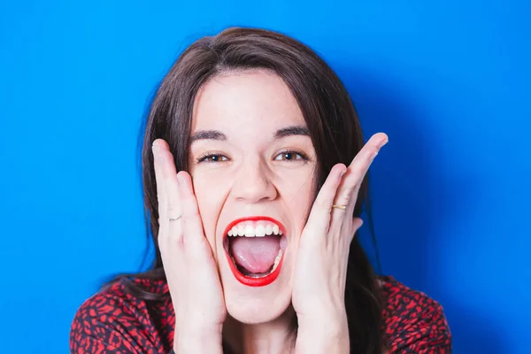 Portrait of positive and surprised woman with open mouth and hands in face scream wow isolated over blue background