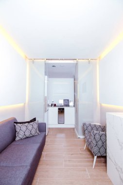 Kanepe, koltuk, duvarlarda beyaz paneller ve koridorun sonunda küçük bir mutfak olan bekleme odası..