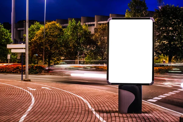Şehirde geceleri boş tuval ekranlı dikey reklam panosu modeli ve hareket halindeki trafik arabaları.