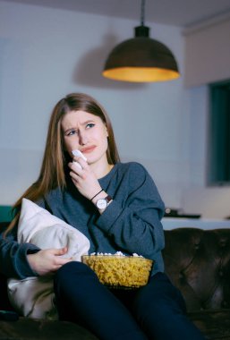 Genç bir kadın evde televizyonda hüzünlü bir film izlerken ağlıyor. Bir kase patlamış mısırla kanepede oturuyor..