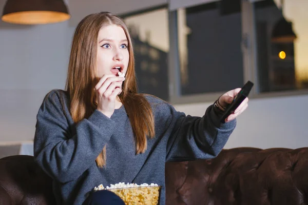 Surprised young beautiful woman watching tv at home while eating popcorn and holding remote control.