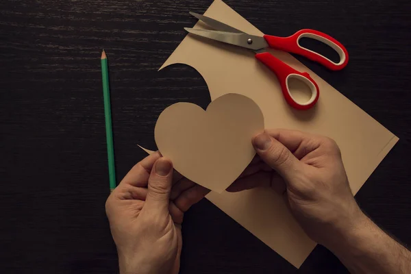 Carved heart in the hands. On a wooden table are scissors, a sheet of cardboard, and a pencil.