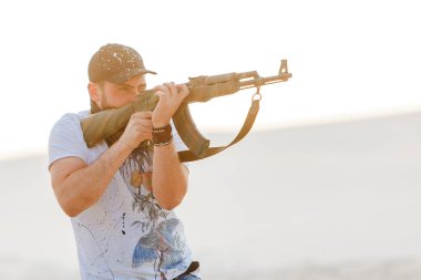 Kızgın adam çöl şapkası bandana kurukafa tişörtü Kalaşnikov silah makineli tüfek gün batımı beyaz kum Barkhan