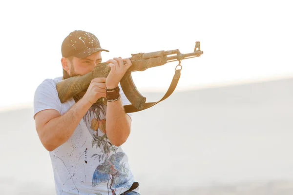 愤怒的男人惹恼了站在那里的沙漠帽乐队骷髅T恤衫卡拉什尼科夫武器机枪夕阳白沙巴汗 — 图库照片