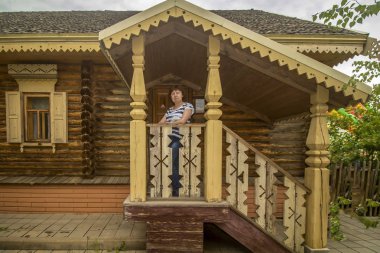Çizgili tişörtlü ve kot pantolonlu bir kadın verandada duruyor ahşap bir evin arka planında.