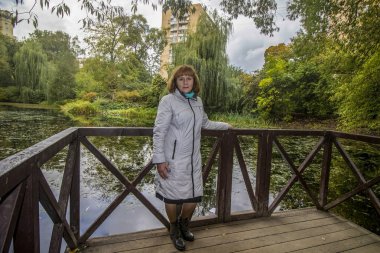 Hafif pelerinli bir bayan, bir köprünün üzerinde, bir gölün ve ağaçların arkasında duruyor.