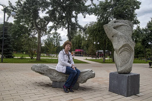 Beyaz ceketli kadın taş parktaki bankta oturuyor.