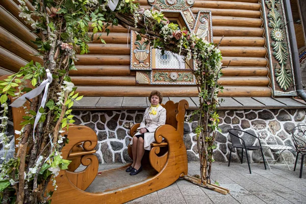 Beyaz pelerinli ve sarı eşarplı bir kadın parktaki ahşap bankta oturuyor.