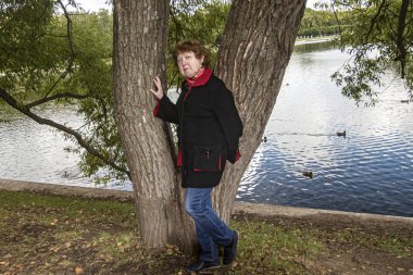Sonbahar. Kırmızı yakalı, siyah ceketli bir bayan, yüzen ördekli bir gölün arka planında bir ağacın yanında duruyor.