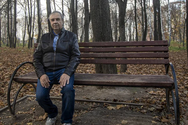 Sonbahar. Deri ceketli ve kot pantolonlu bir adam bankta oturuyor.