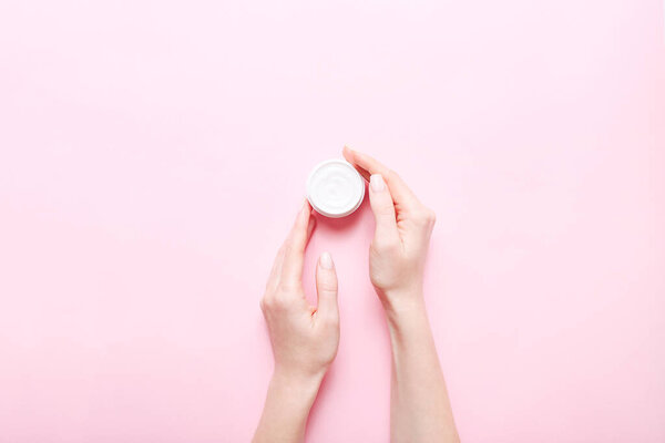 Open jar with cream for skin care in female hands on pink background.
