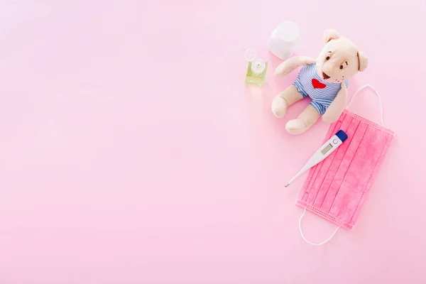 Védőmaszk medve játékkal, hőmérővel és kézfertőtlenítővel rózsaszín háttérrel. Az iskolák karanténjának koncepciója, otthoni oktatás — Stock Fotó