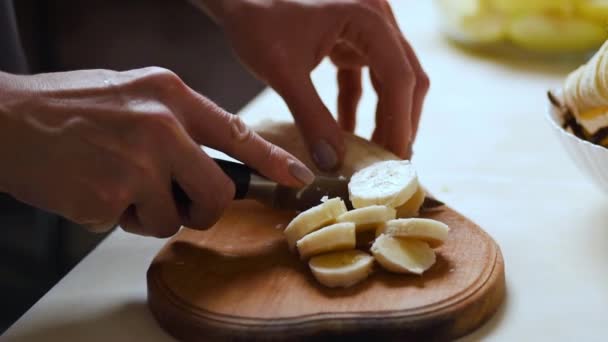 Manos femeninas cortando por cuchillo fruta de plátano sobre tabla de madera. Primer plano cocinera cortar plátano a rebanadas en la tabla de la cocina — Vídeos de Stock