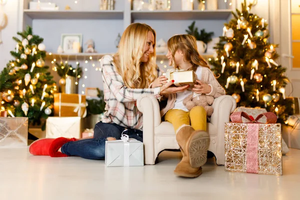 New Years picture of mother giving gift to daughter sitting on chair against backdrop of scenery, Christmas tree
