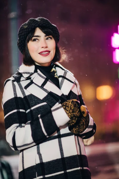 Glückliche Frau in kariertem Mantel und schwarzem Hut beim Abendspaziergang auf verschwommenem rosa Hintergrund — Stockfoto