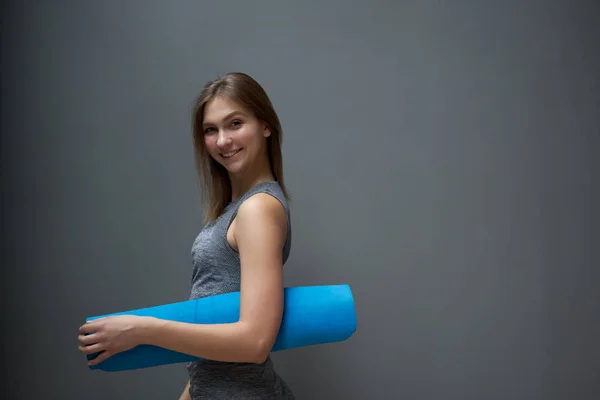 Šťastná sportovkyně s modrým kobercem v rukou na pozadí šedé stěny — Stock fotografie