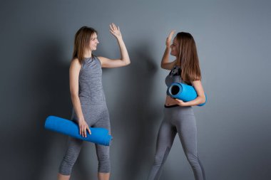 Two sporty women doing handshake while standing with twisted blue rugs against gray empty wall clipart