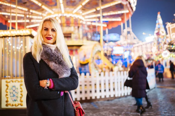 Bild einer jungen Frau im Mantel mit Pelzkragen im Park auf dem Hintergrund des Karussells — Stockfoto