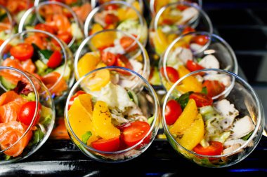 Kırmızı balık, domates, salatalık, yeşillik, et, portakal dolu cam bardaklarla dolu açık büfe masasının üstünde.